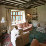 A cozy traditional living room designed by a interior designer in kent , featuring original dark wood ceiling beams, a built-in library wall, and a bright window seat overlooking a garden.