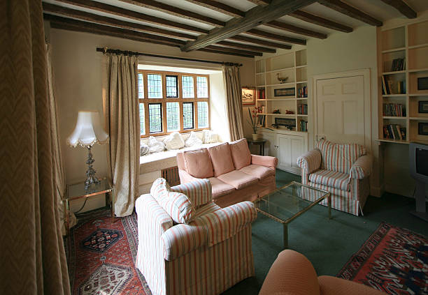 A cozy traditional living room designed by a interior designer in kent , featuring original dark wood ceiling beams, a built-in library wall, and a bright window seat overlooking a garden.