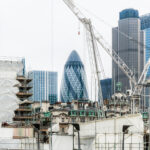 A high-scale urban building site managed by professional construction companies near me, featuring white luffing jib cranes and extensive scaffolding with the iconic Gherkin skyscraper in the background.