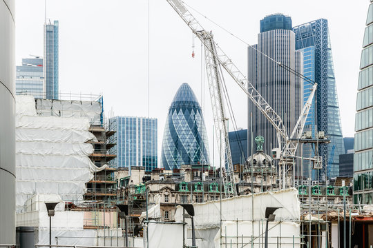 A high-scale urban building site managed by professional construction companies near me, featuring white luffing jib cranes and extensive scaffolding with the iconic Gherkin skyscraper in the background.
